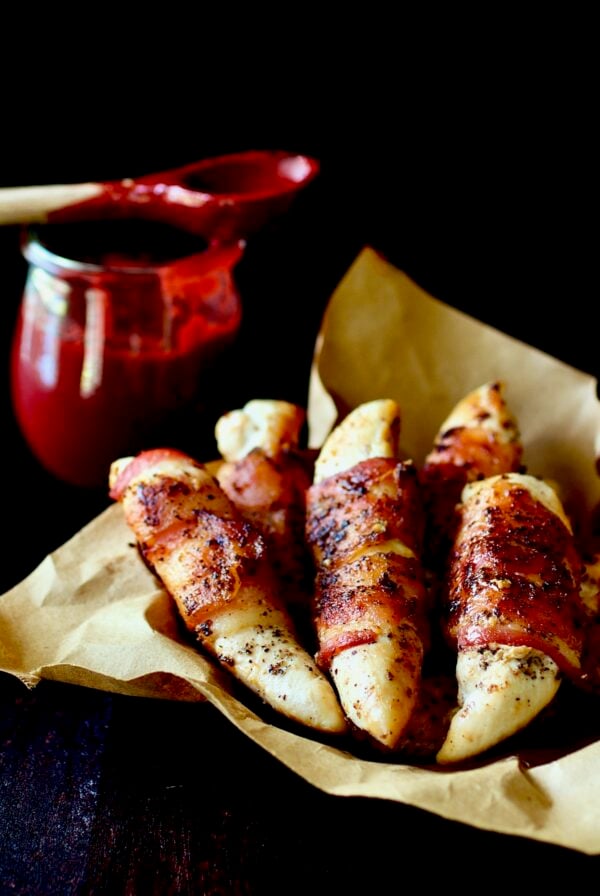 Bacon-Wrapped Chicken Tenders in brown paper lined basket with Raspberry Chipotle Sauce in the background.
