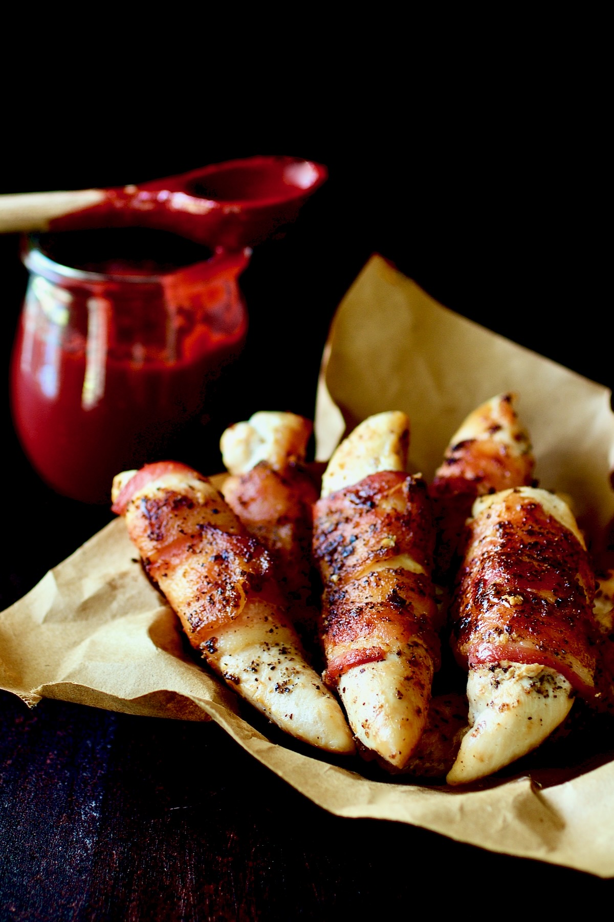Bacon-Wrapped Chicken Tenders in brown paper lined basket with Raspberry Chipotle Sauce in the background.
