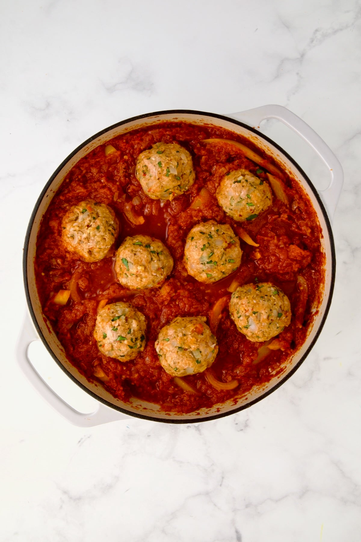 Eight uncooked Harissa Chicken Meatballs placed into the sauce in white enamel cast iron skillet.