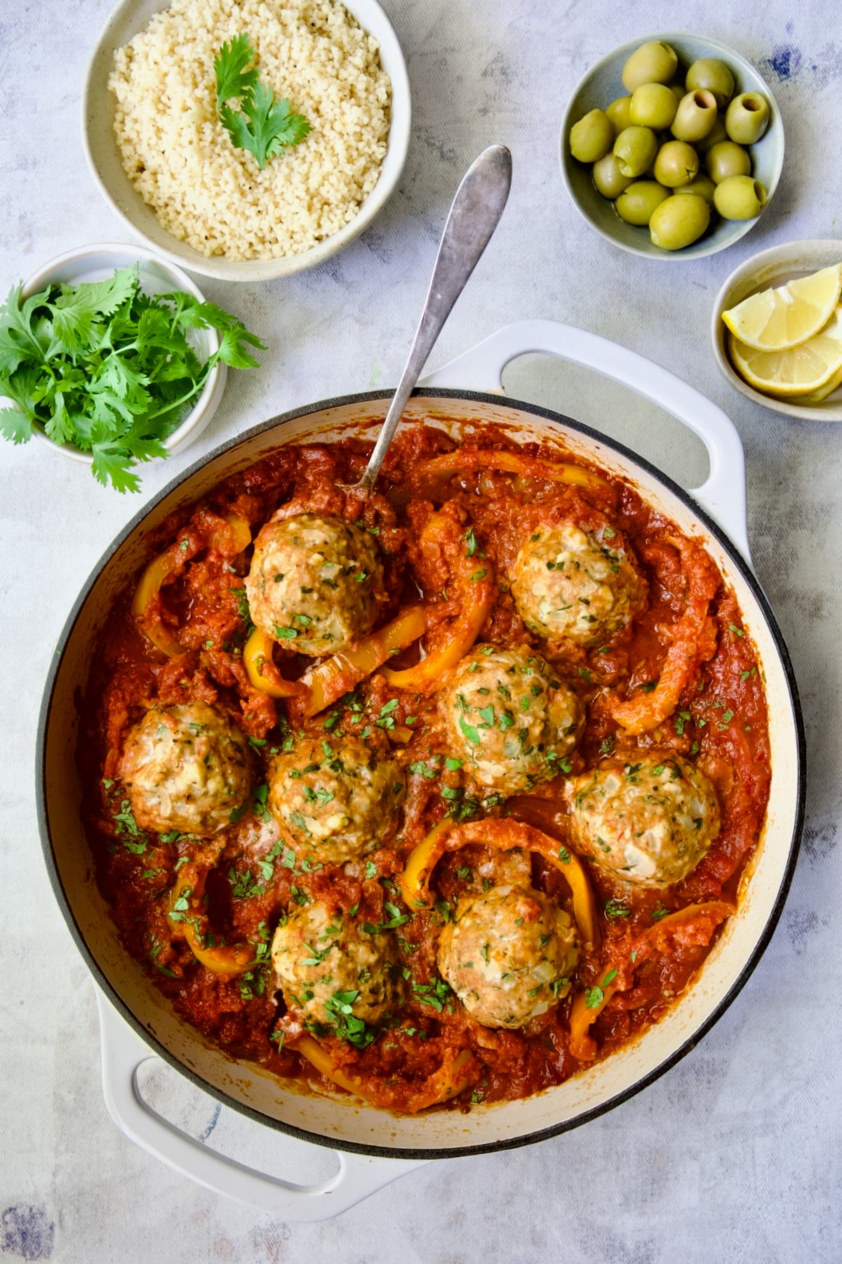 Eight cooked Harissa Chicken Meatballs in the sauce in white enamel cast iron skillet with serving spoon picking one up.