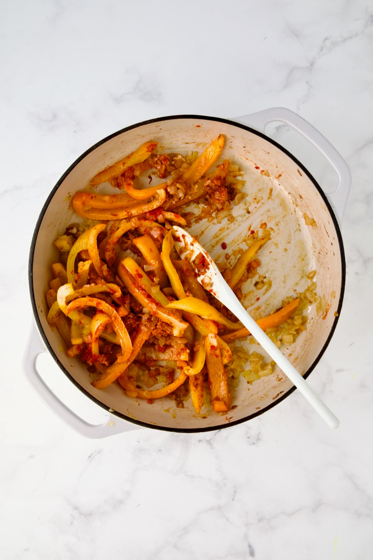 Cooked onions and bell peppers in white enamel cast iron skillet with garlic, cumin, coriander and harissa added being stirred.