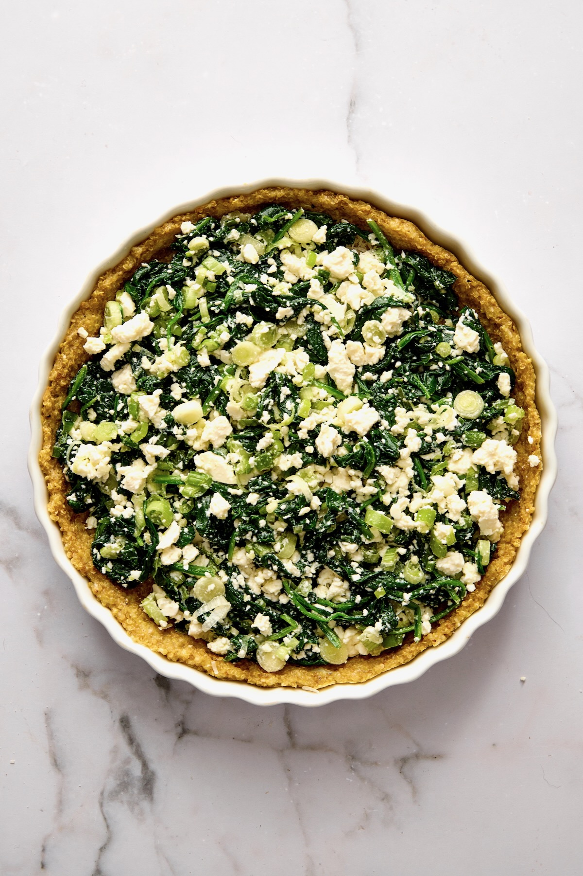 Spinach, scallion and feta cheese in baked quinoa crust in round white baking dish.