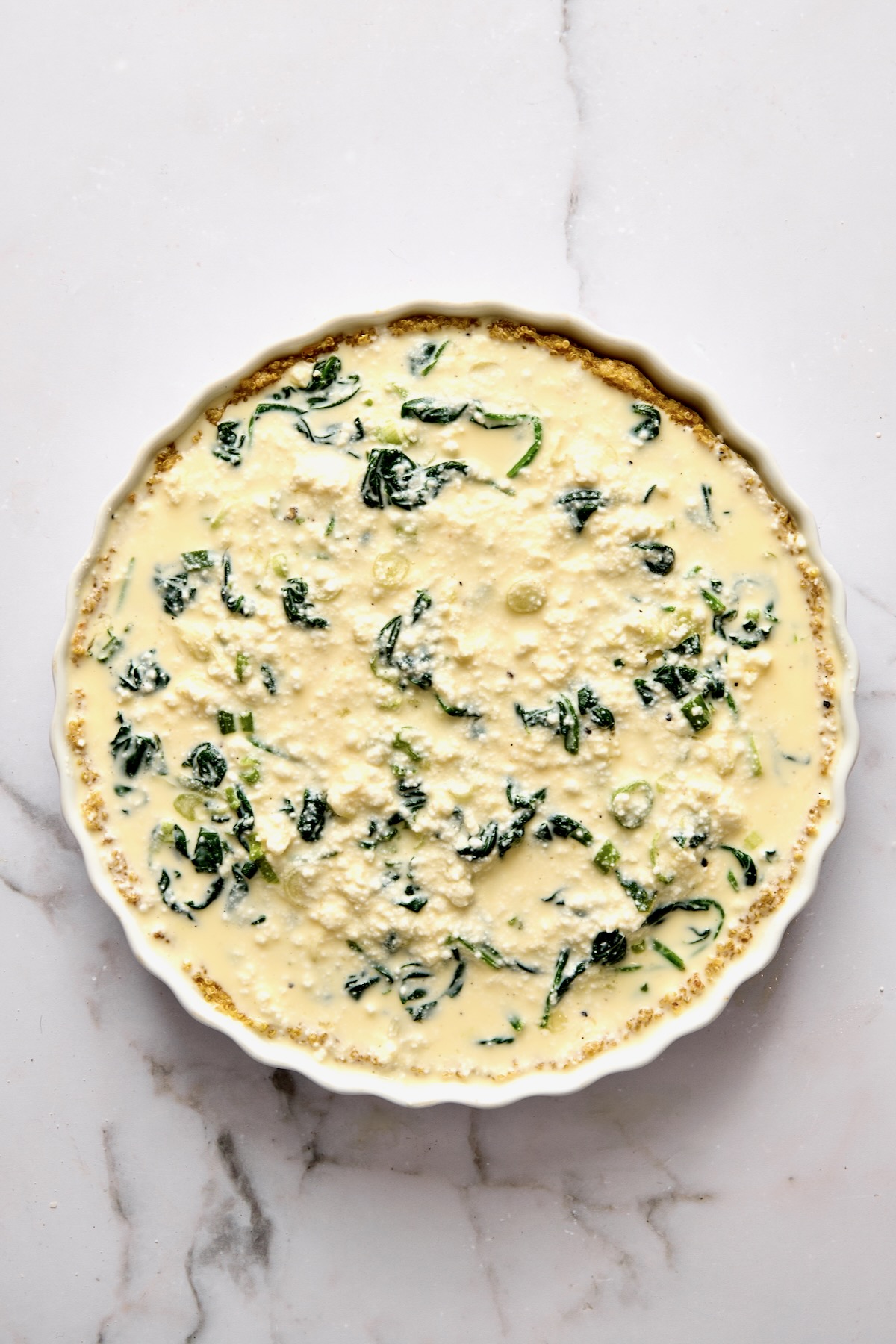 Custard for quiche poured over the other ingredients in round white baking dish.