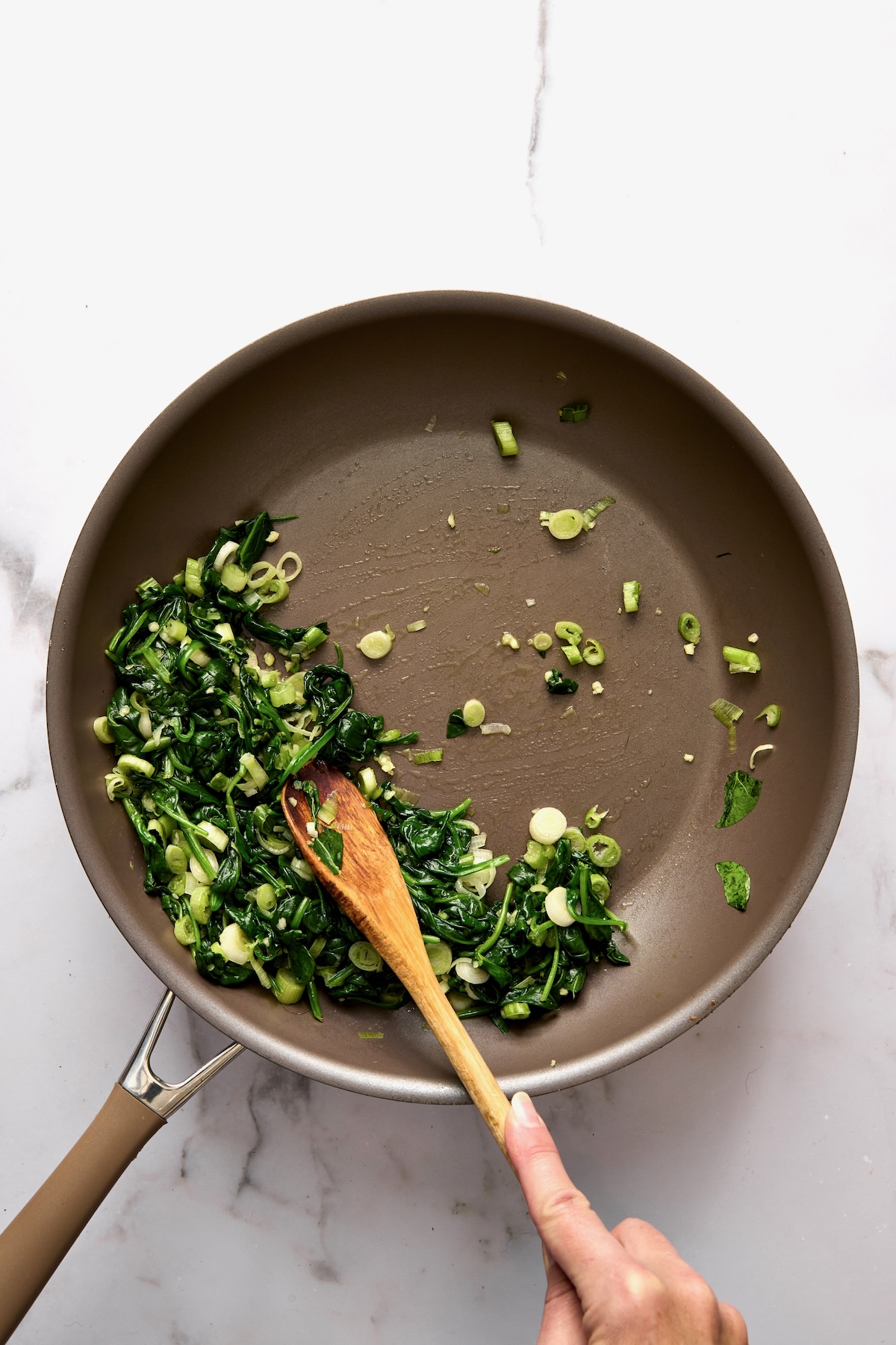 Cooked spinach and scallions in skillet being stirred with wooden spoon.