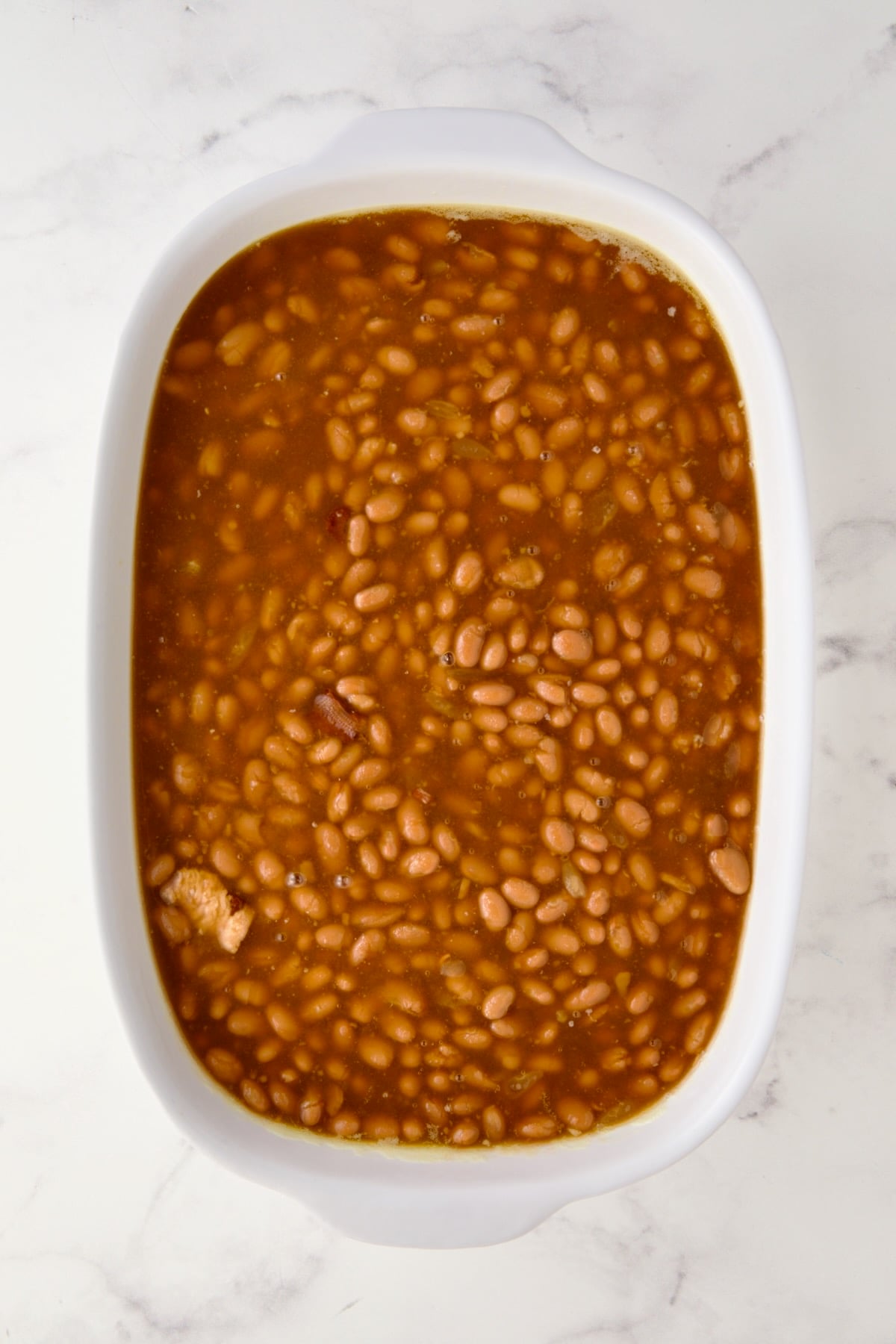 Canned baked beans before other ingredients are added in oval white baking dish.