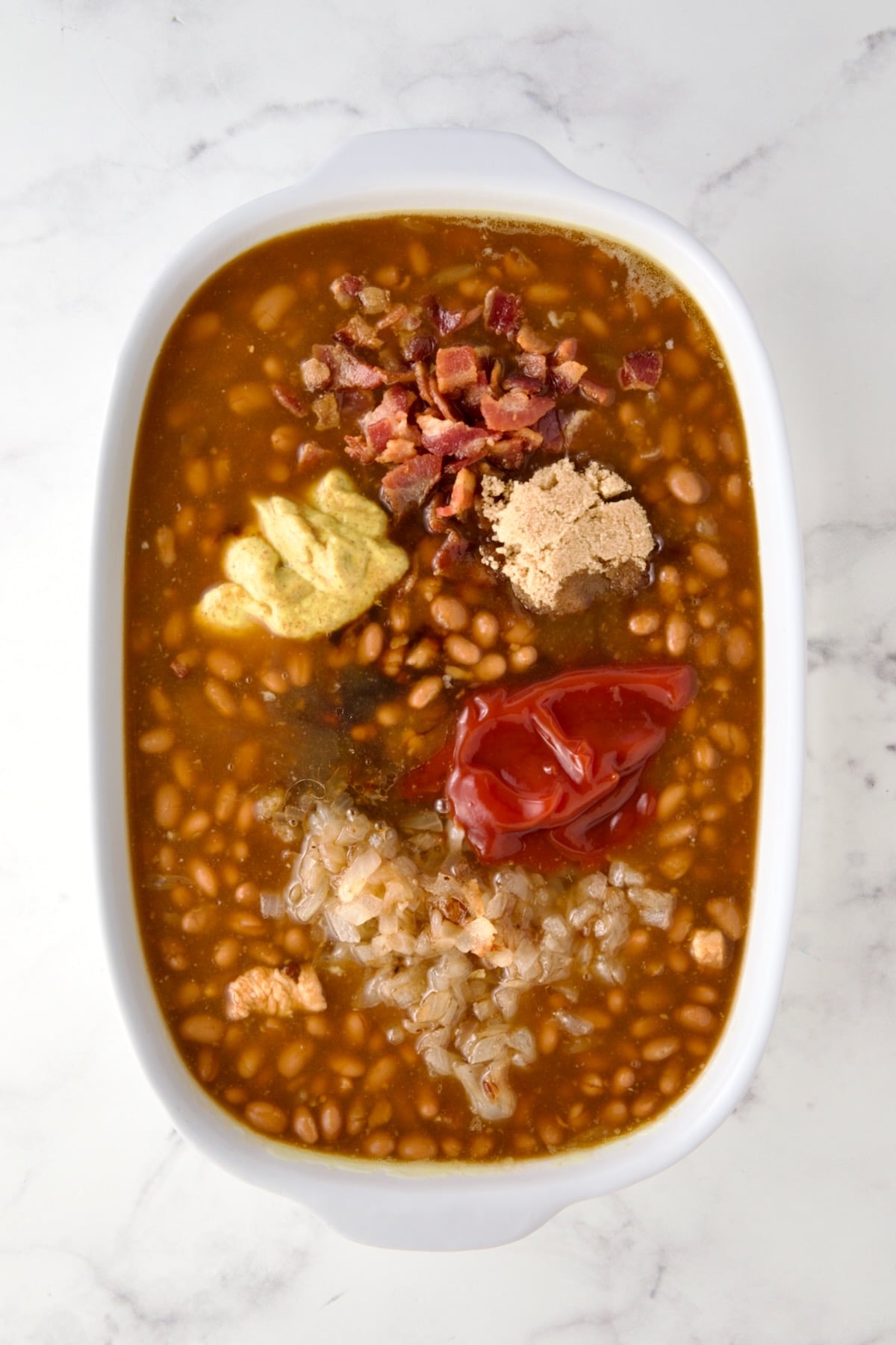 Ingredients added to canned baked beans before being stirred in in oval white baking dish.