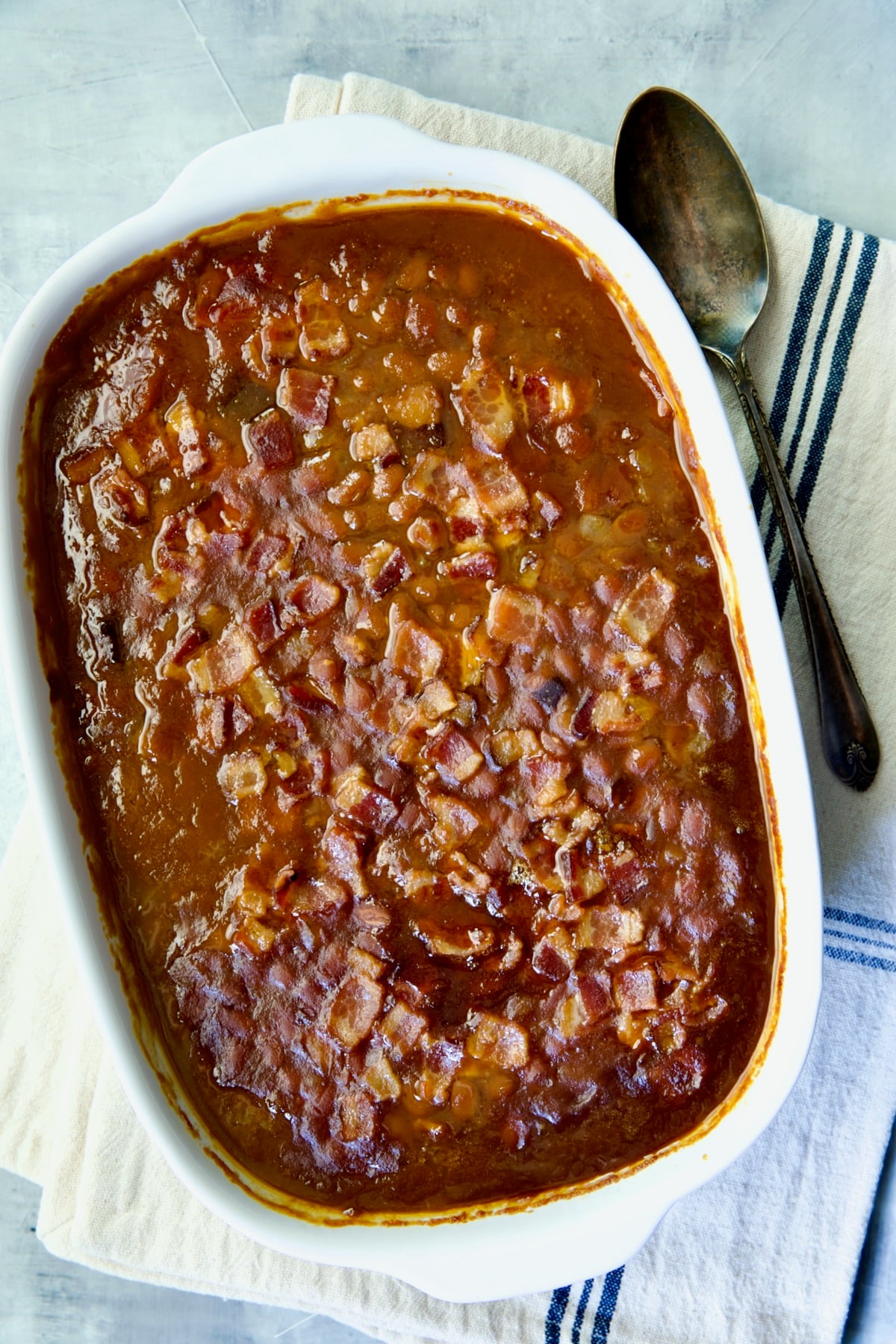 Best Canned Baked Beans Recipe in oval white baking dish with serving spoon on black striped towel.