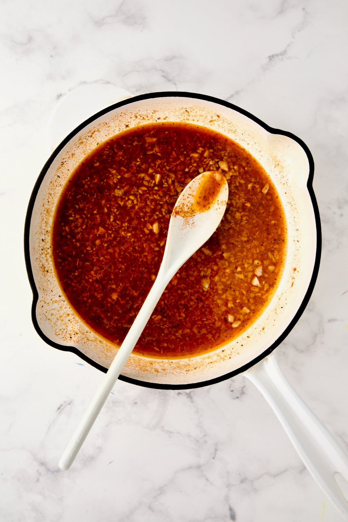 Reduced sauce in white enamel cast iron skillet with white mixing spoon.