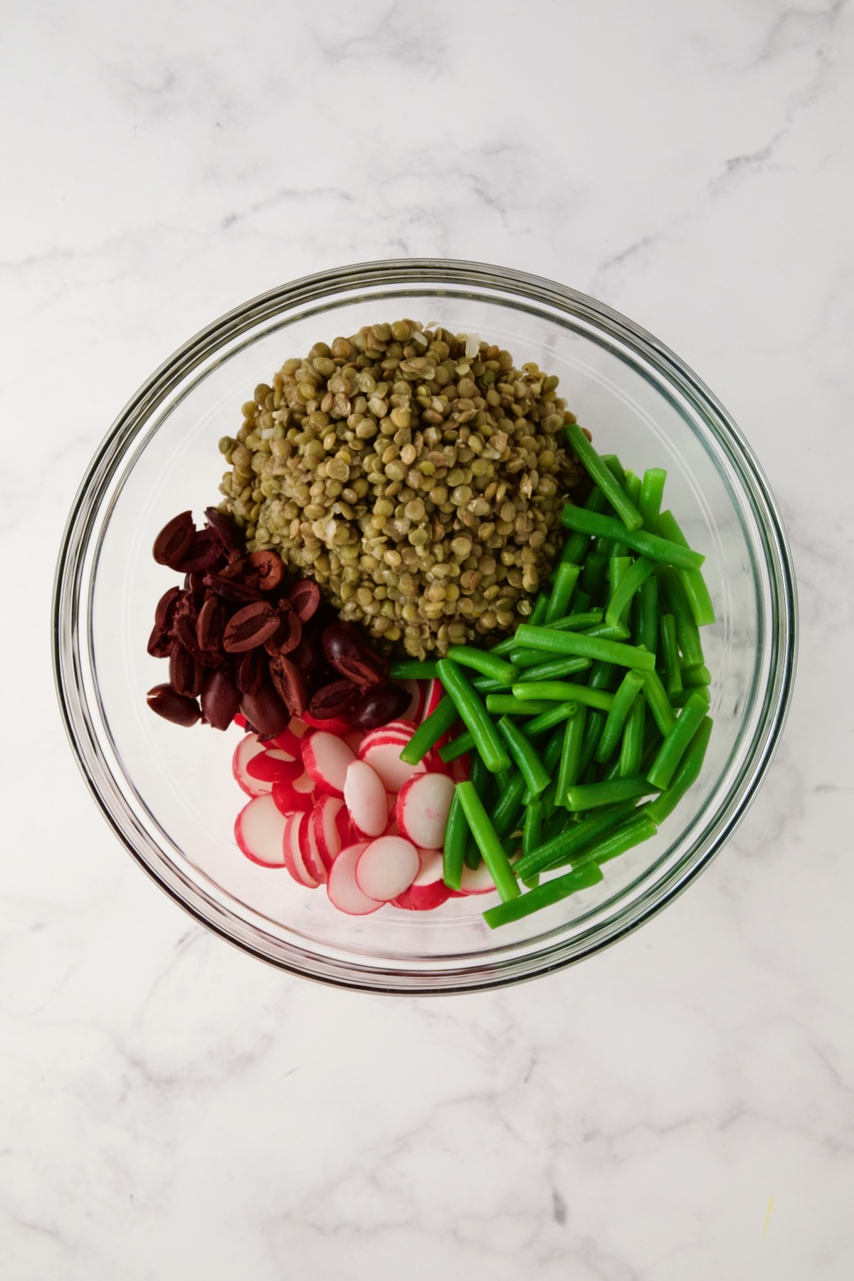 Cooked lentils, blanched green beans, sliced radishes and halved Kalamata olives in glass bowl before being mixed together.