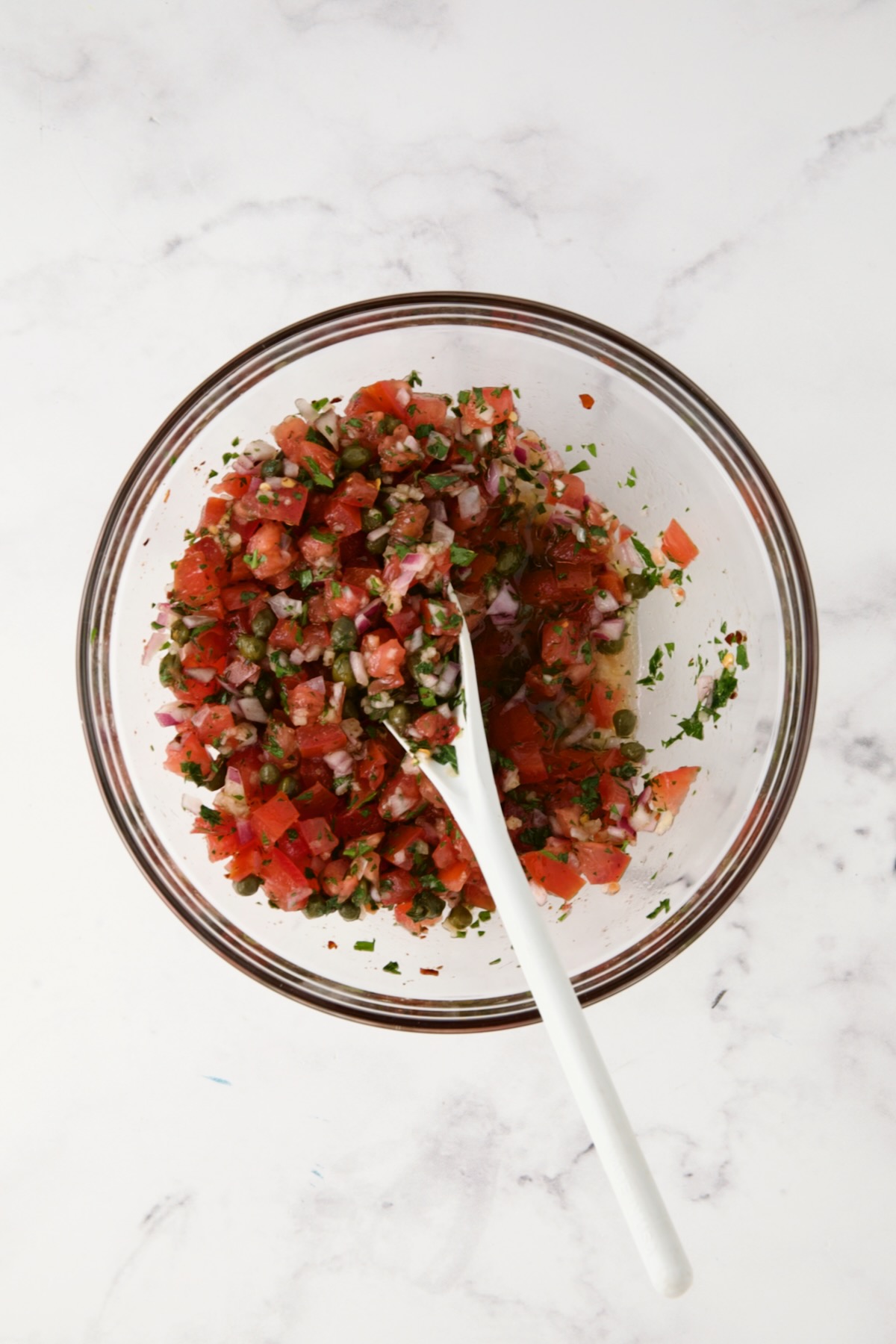 Tomato Caper Relish in glass bowl being mixed with white spoon.