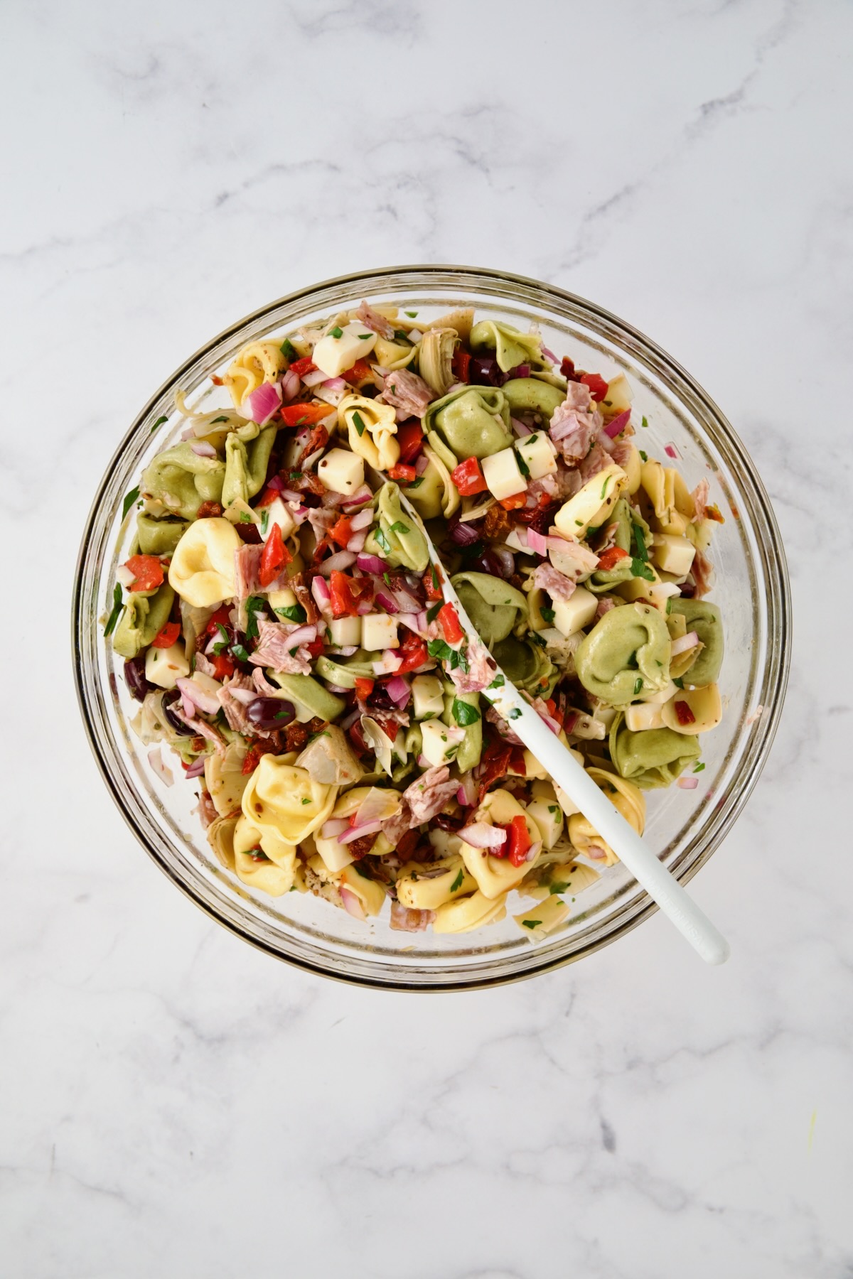 Ingredients for tortellini salad in glass mixing bowl after being combined with white mixing spoon.