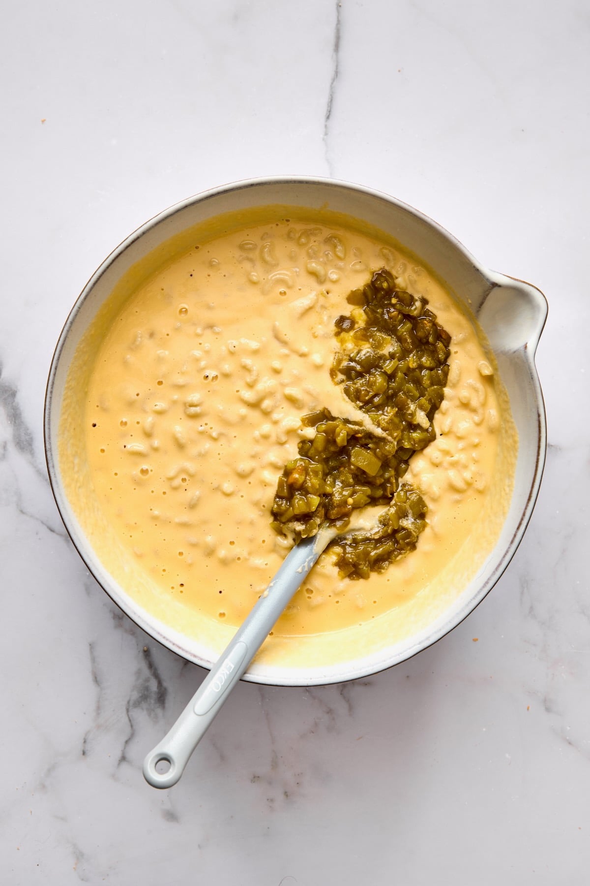 Green chiles added to cheese sauce and macaroni in white mixing bowl.
