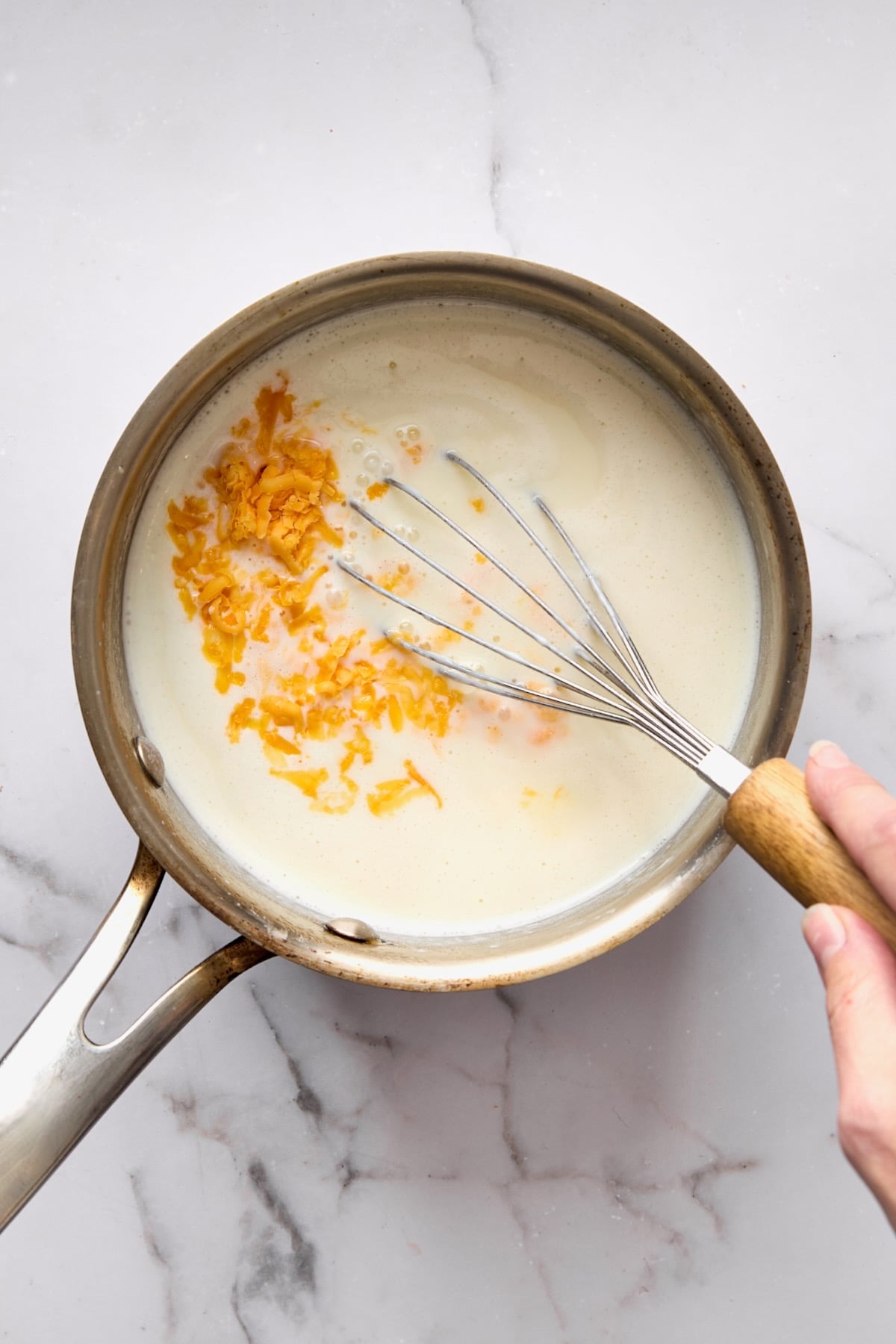 Cheddar cheese added to thickened milk being whisked in stainless steel saucepan.