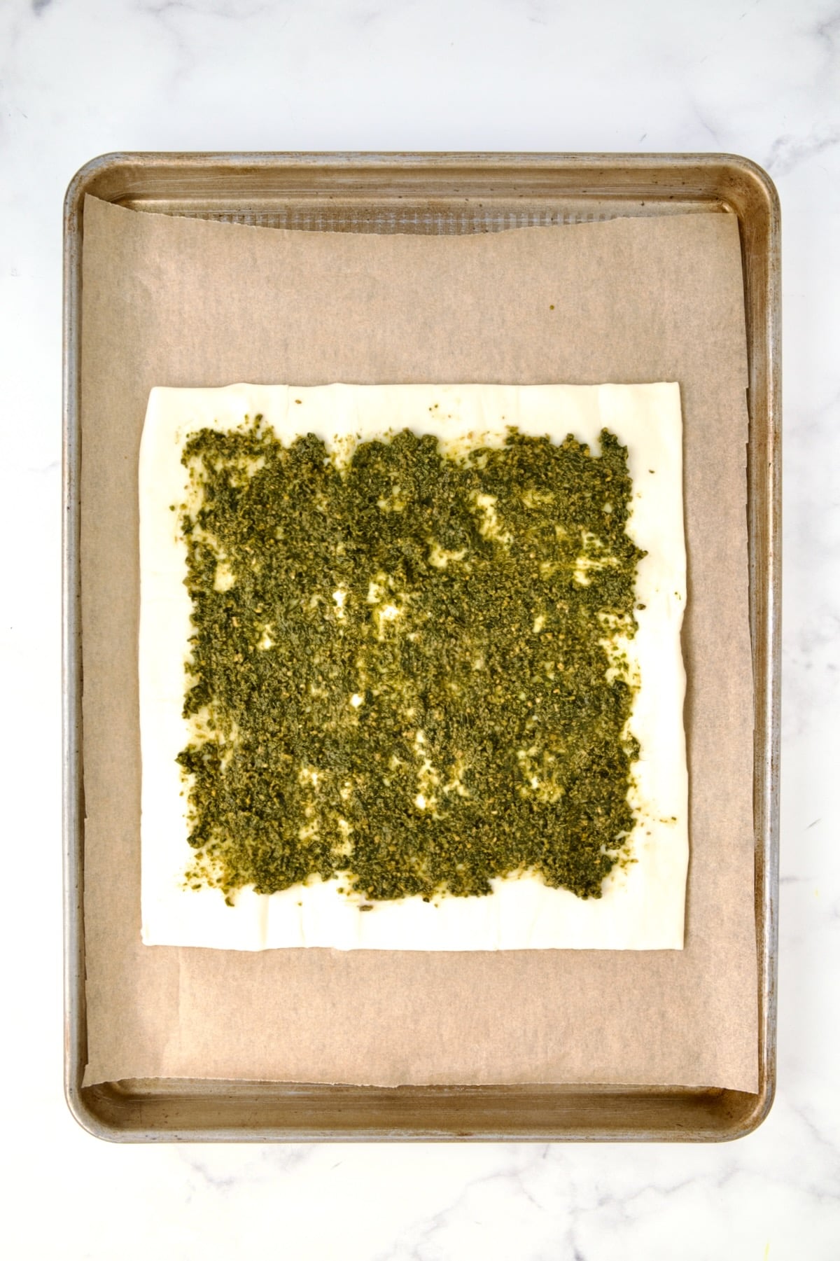 Puff pastry piece on parchment paper-lined rimmed baking sheet with pesto spread onto it.