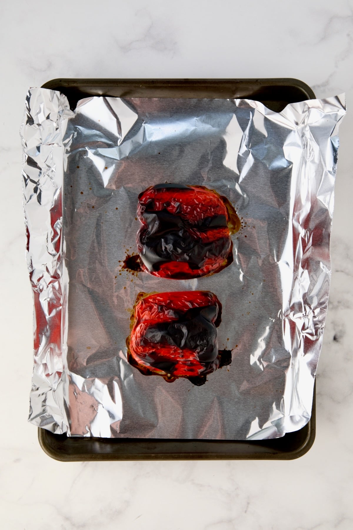 Two red bell pepper halves on aluminum foil-lined rimmed sheet pan after being roasted.