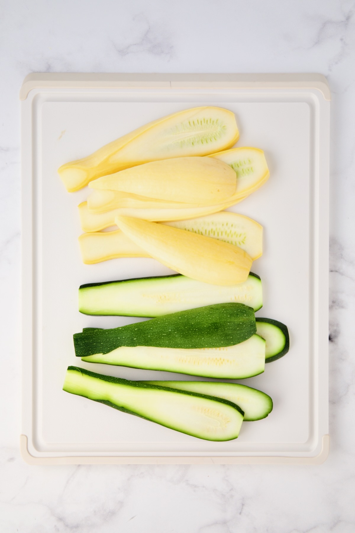 Sliced yellow squash and zucchini on white plastic cutting board.