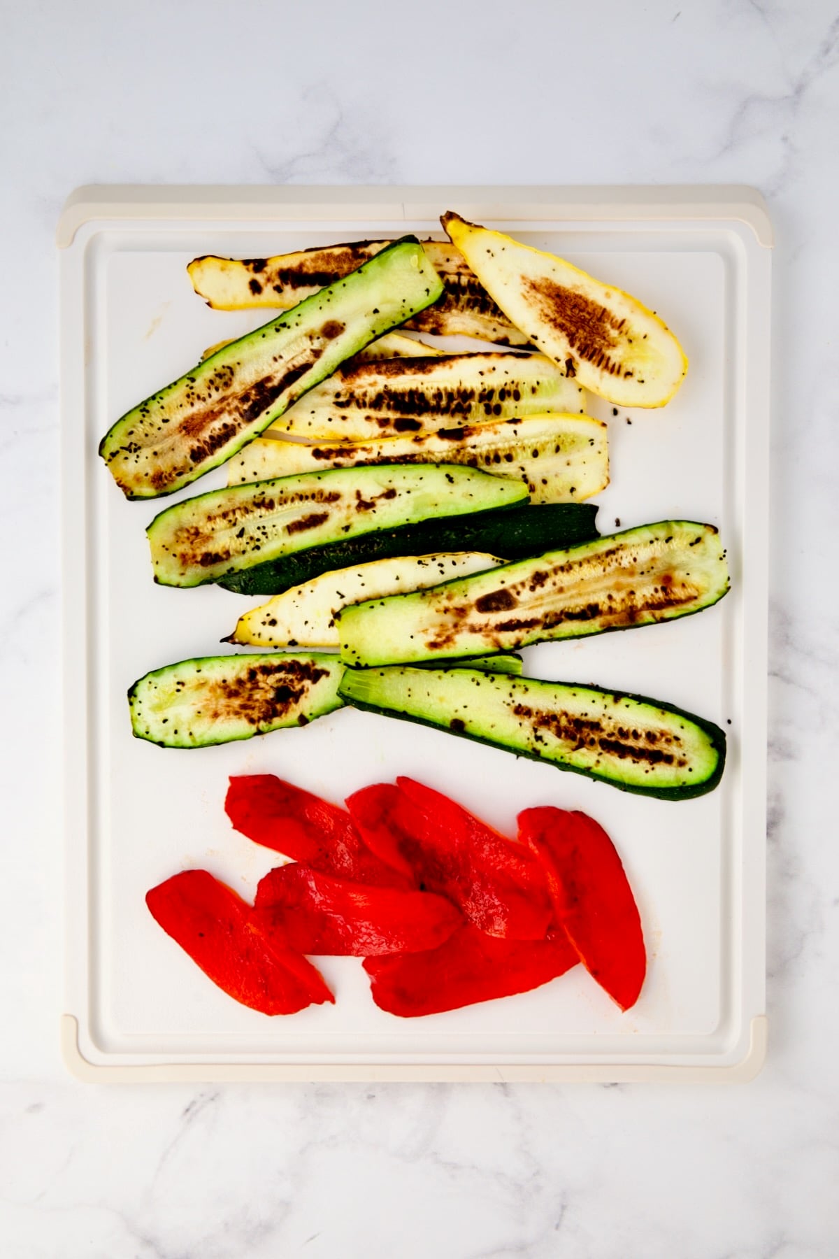 Charred yellow squash, zucchini and roasted red bell pepper slices on white plastic cutting board.