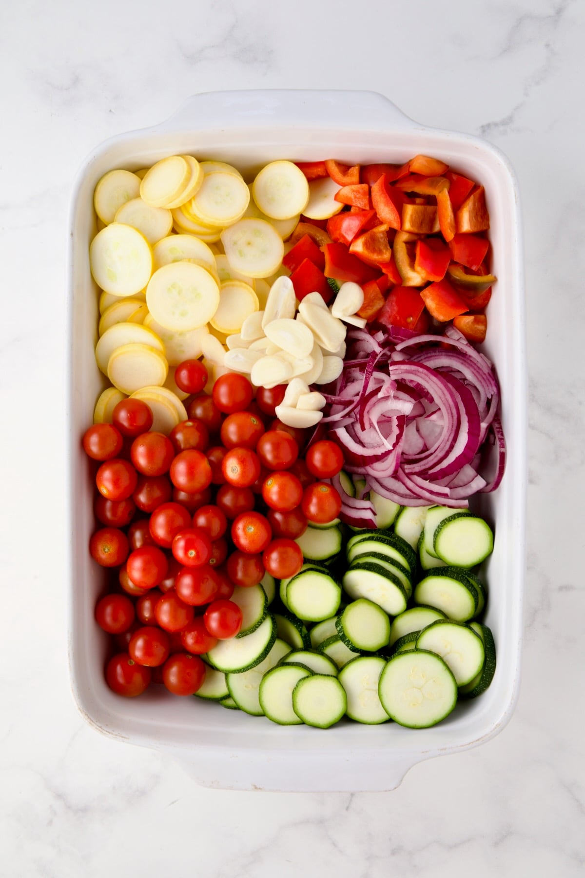 Uncooked cherry tomatoes, yellow squash, zucchini, red bell pepper, red onion and sliced garlic in white rectangular baking dish before being combined.