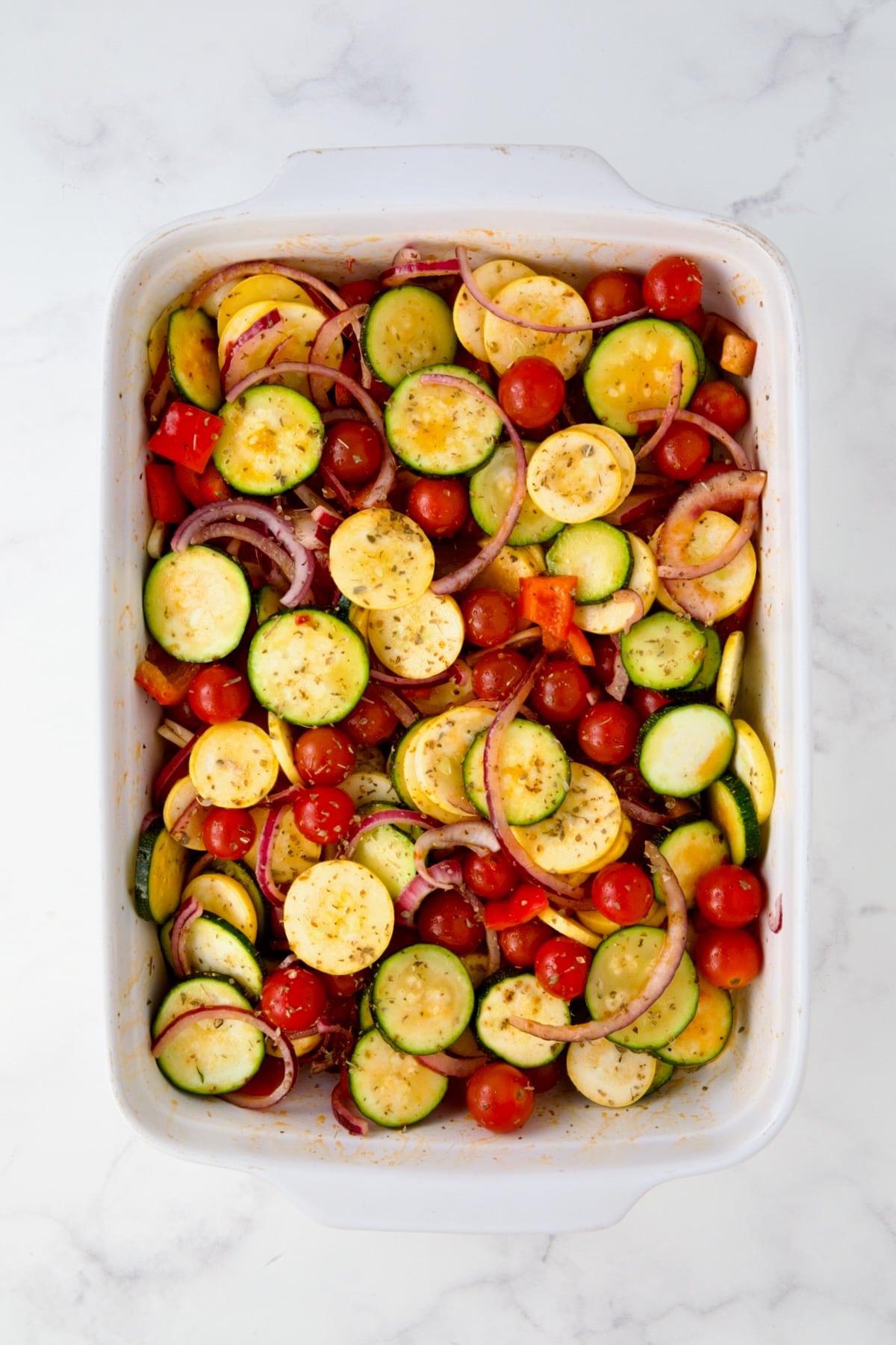 Uncooked cherry tomatoes, yellow squash, zucchini, red bell pepper, red onion and sliced garlic in white rectangular baking dish after being combined.