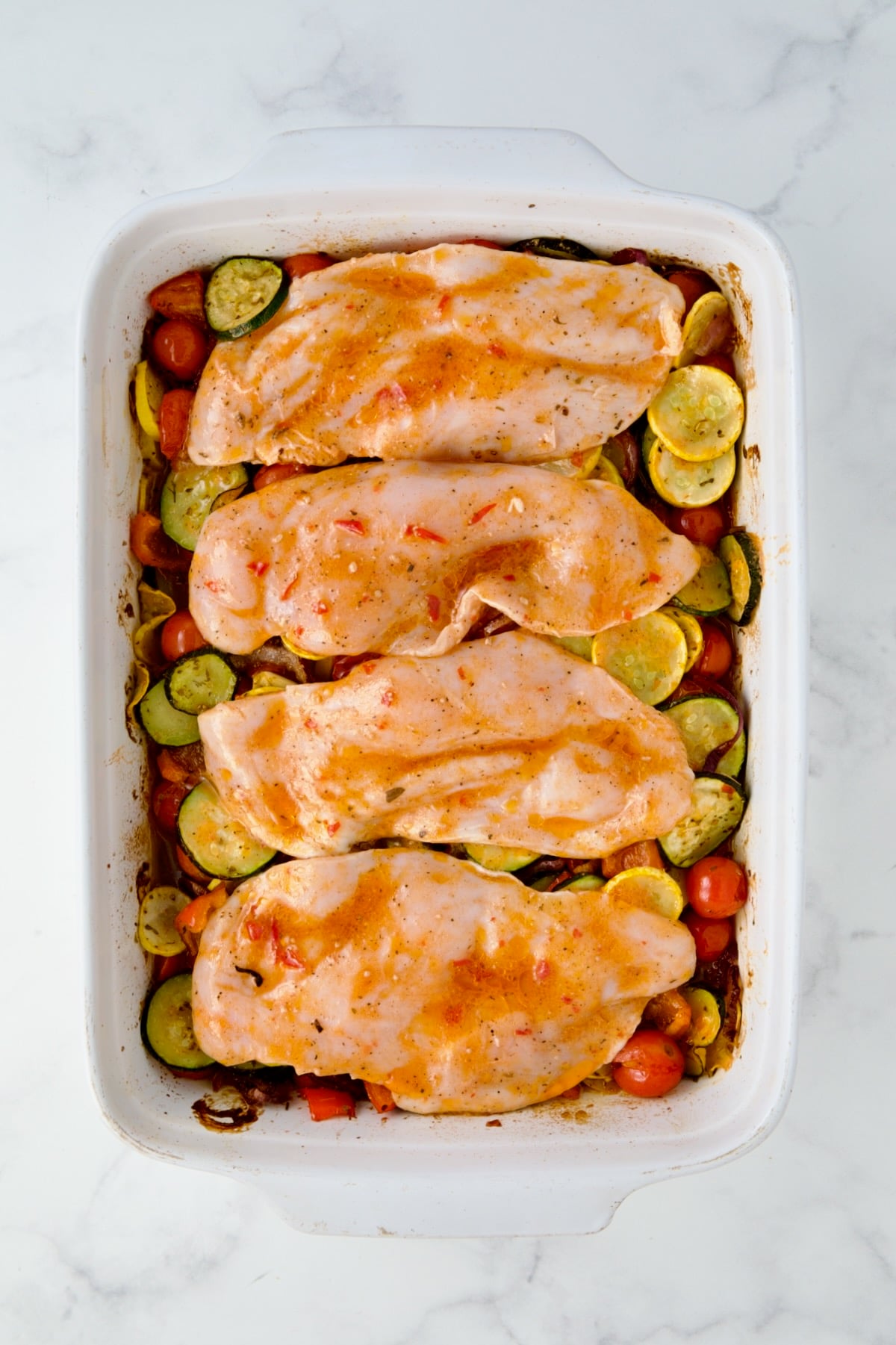 Four uncooked chicken cutlets placed over semi-cooked vegetables in white rectangular baking dish.