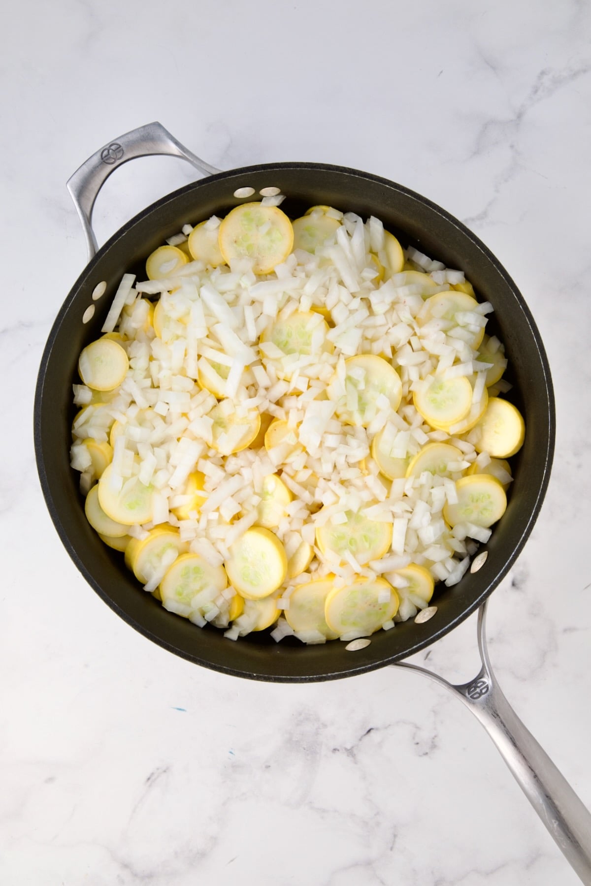 Uncooked sliced yellow squash and chopped onion in black skillet.