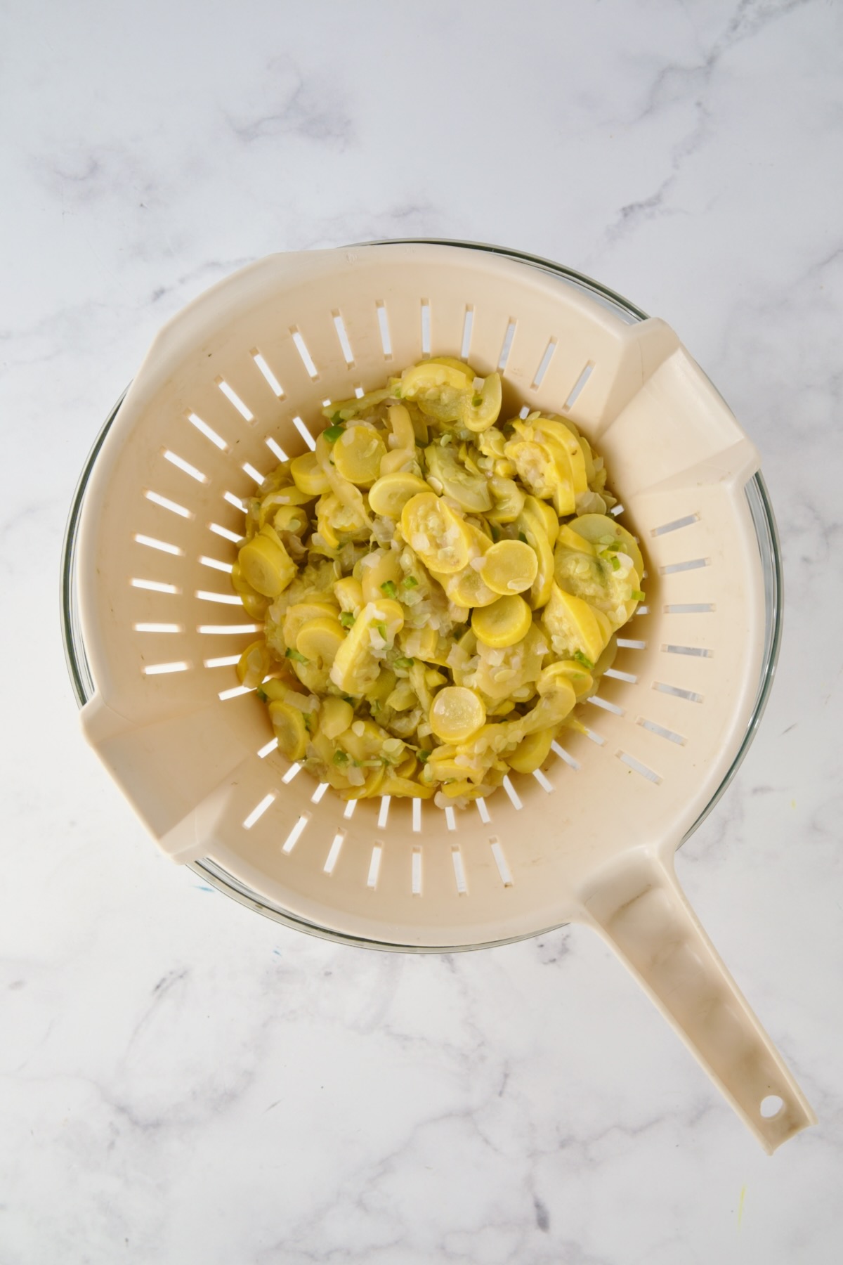 Cooked yellow squash, onion and jalapeno in beige colander set over a glass bowl.