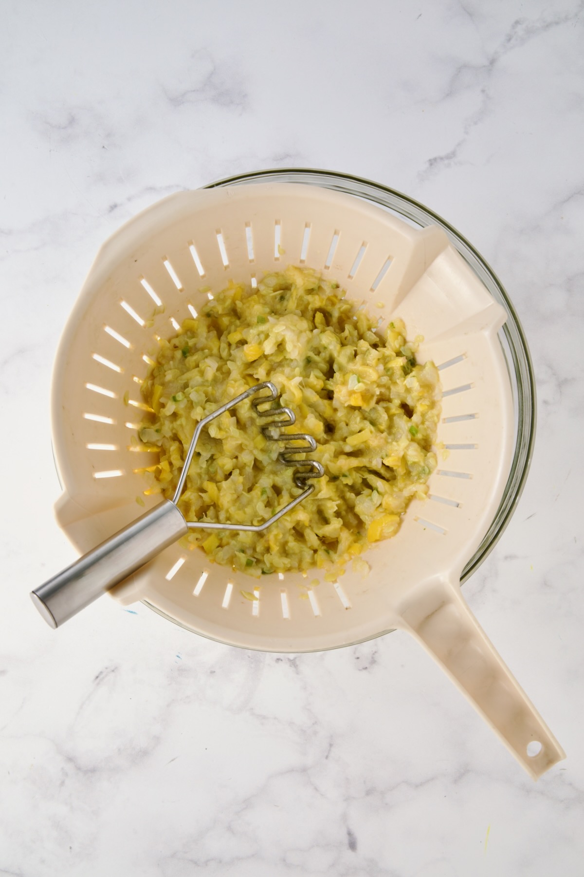 Cooked mashed yellow squash, onion and jalapeno in beige colander set over a glass bowl with potato masher.