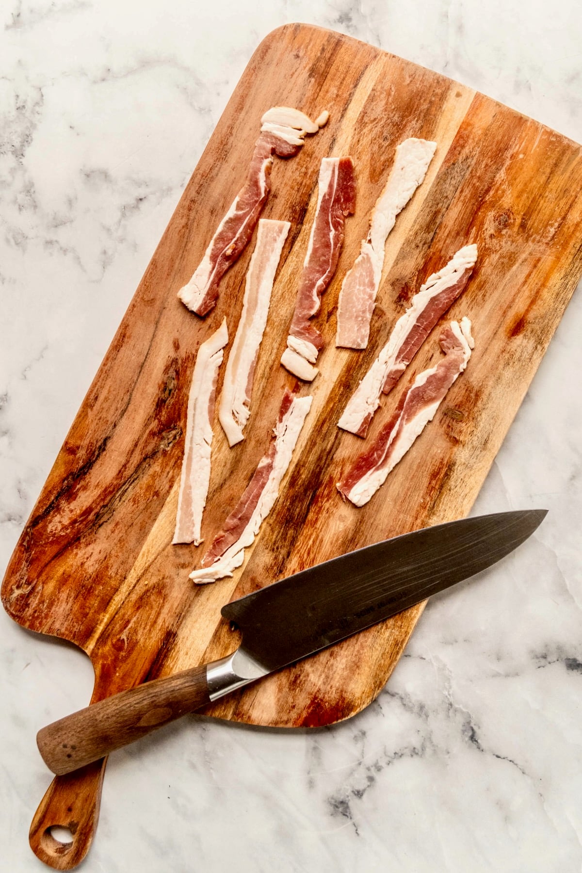 Small strips of uncooked bacon on wooden cutting board with chef's knife.