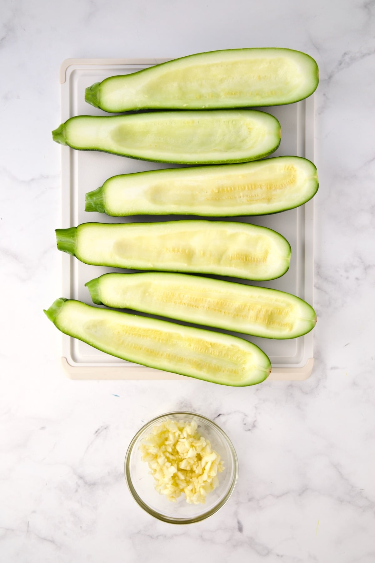 Six hollowed out zucchini halves on white plastic cutting board with the pulp in a small glass bowl.