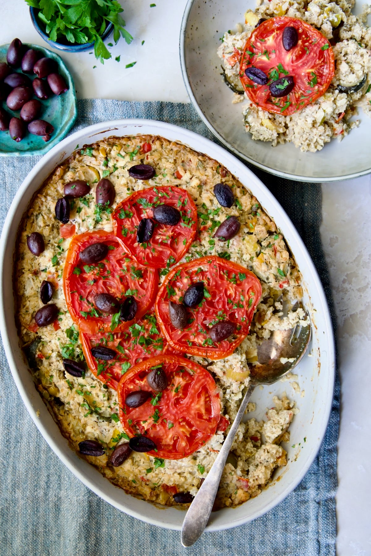 Greek Vegetable Casserole Recipe in oval baking dish with serving spoon and some in a dinner bowl above it.