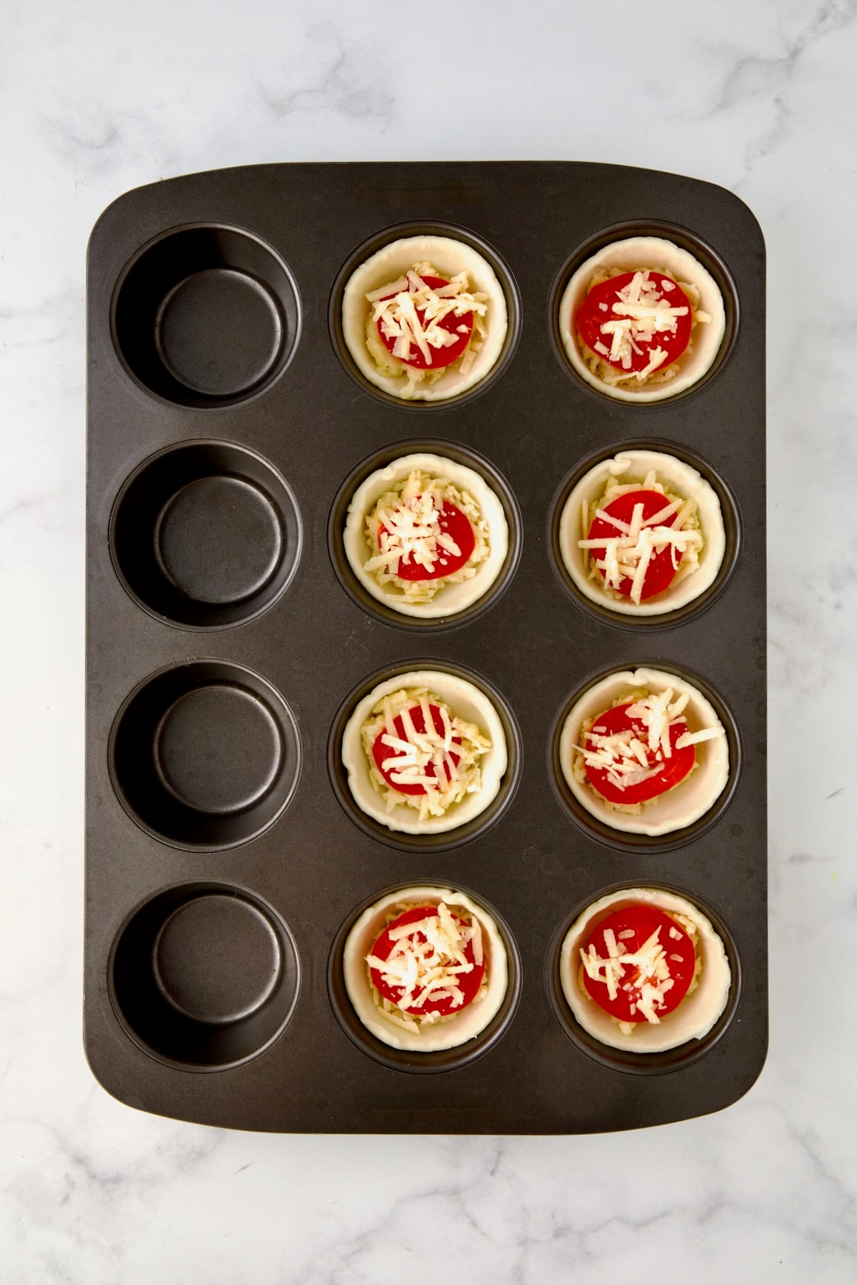 Cheese sprinkled over the top of tomatoes in a 12-cup metal muffin pan.