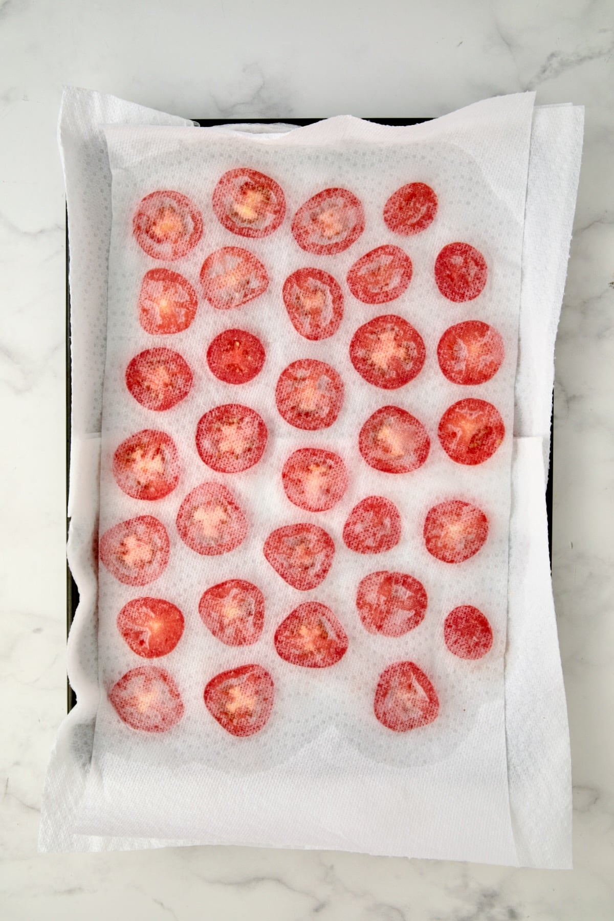 Sliced plum tomatoes on paper towel lined rimmed baking sheet with a layer of paper towels over them.
