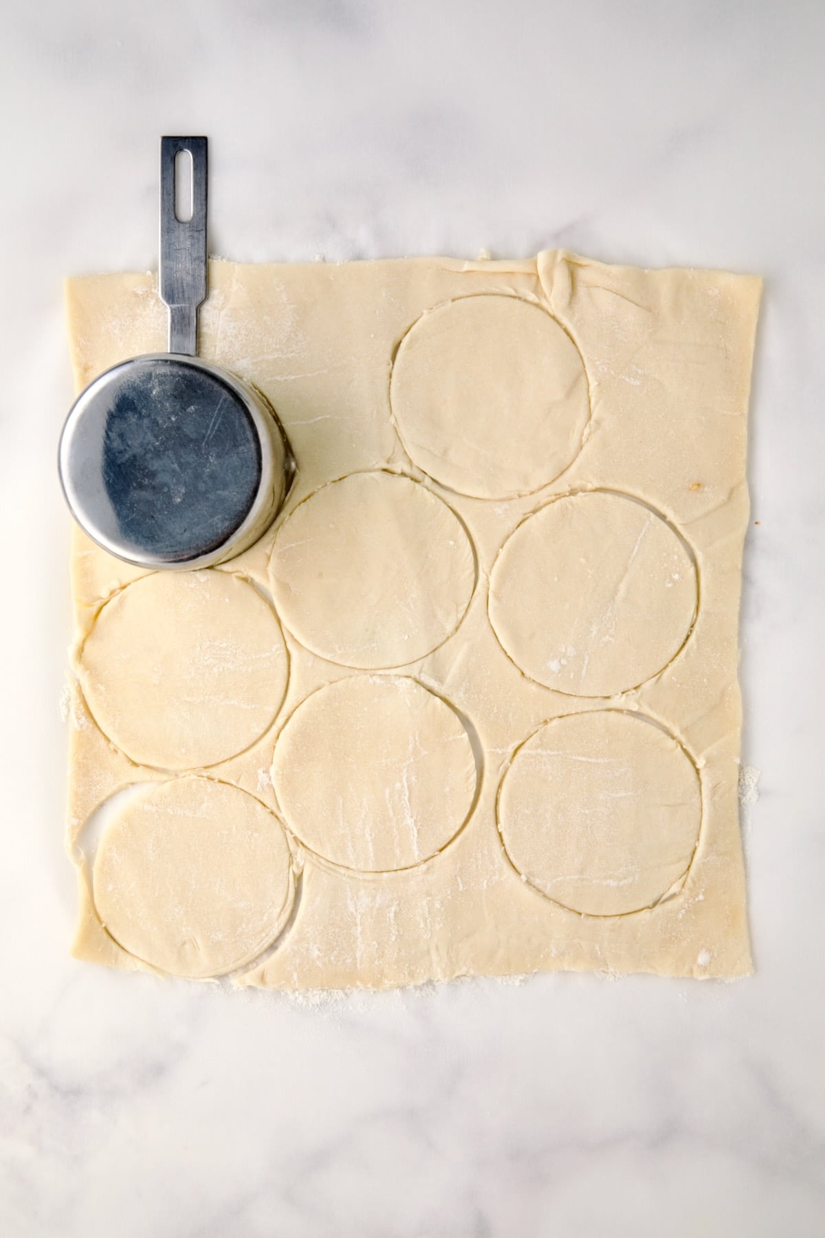 Sheet of puff pastry with round pieces cut by a stainless steel measuring cup.