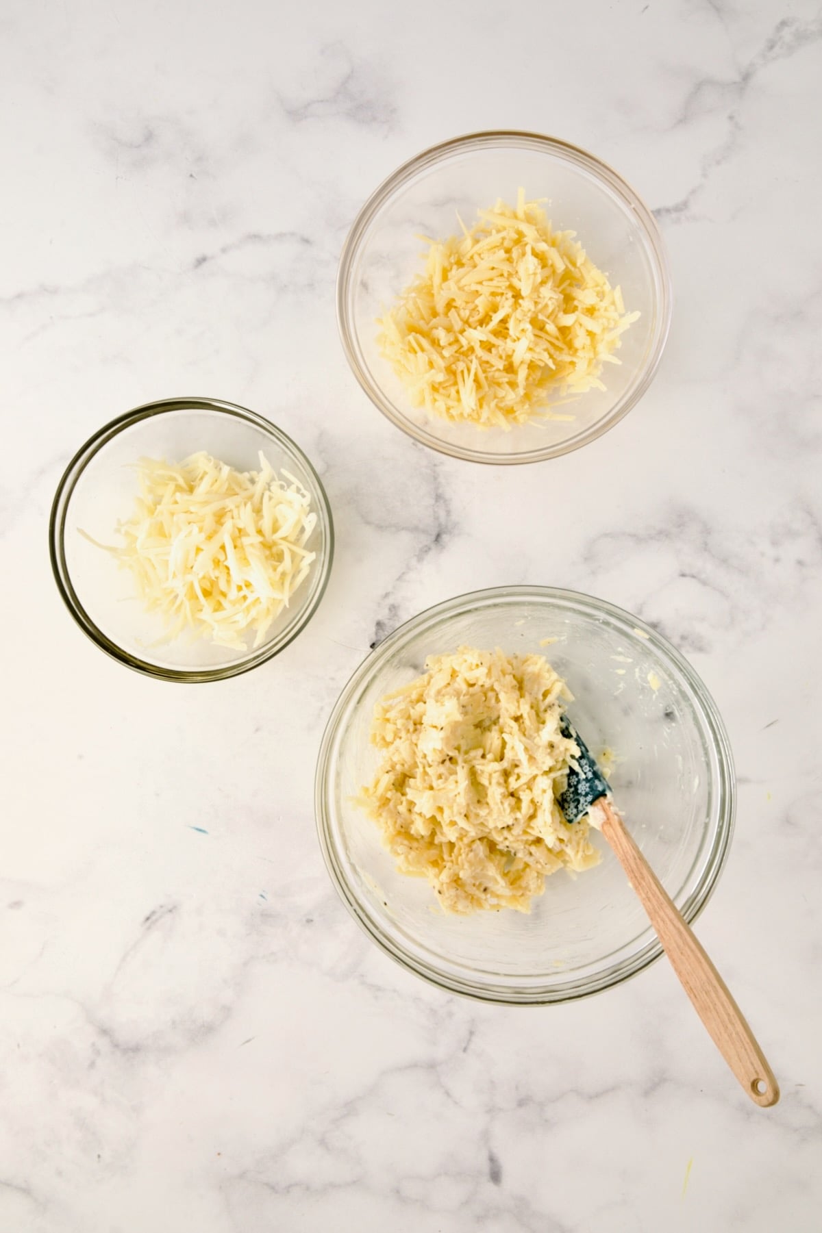 Three glass bowls; one with cheese and mayonnaise combined with small spatula and two bowls of grated cheese.