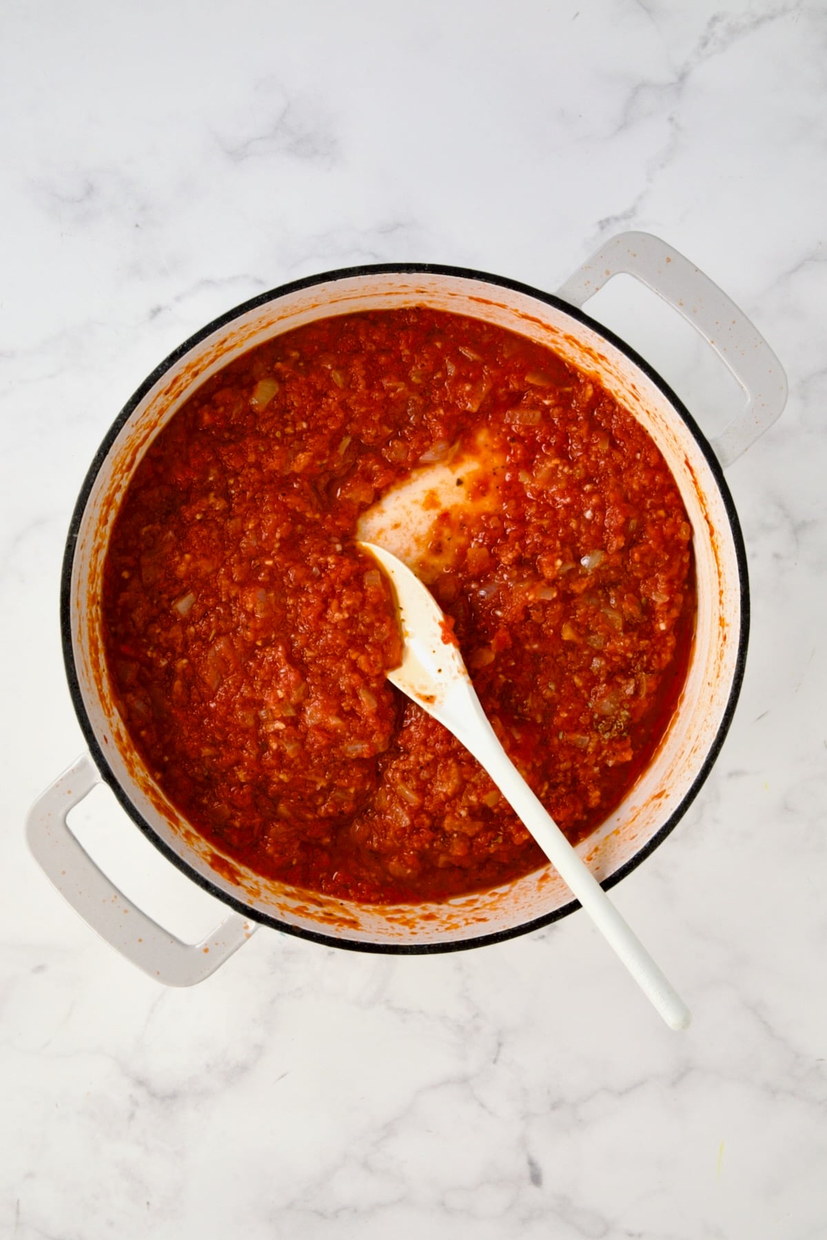 White plastic spoon stirring tomato sauce in white enamel cast iron skillet.