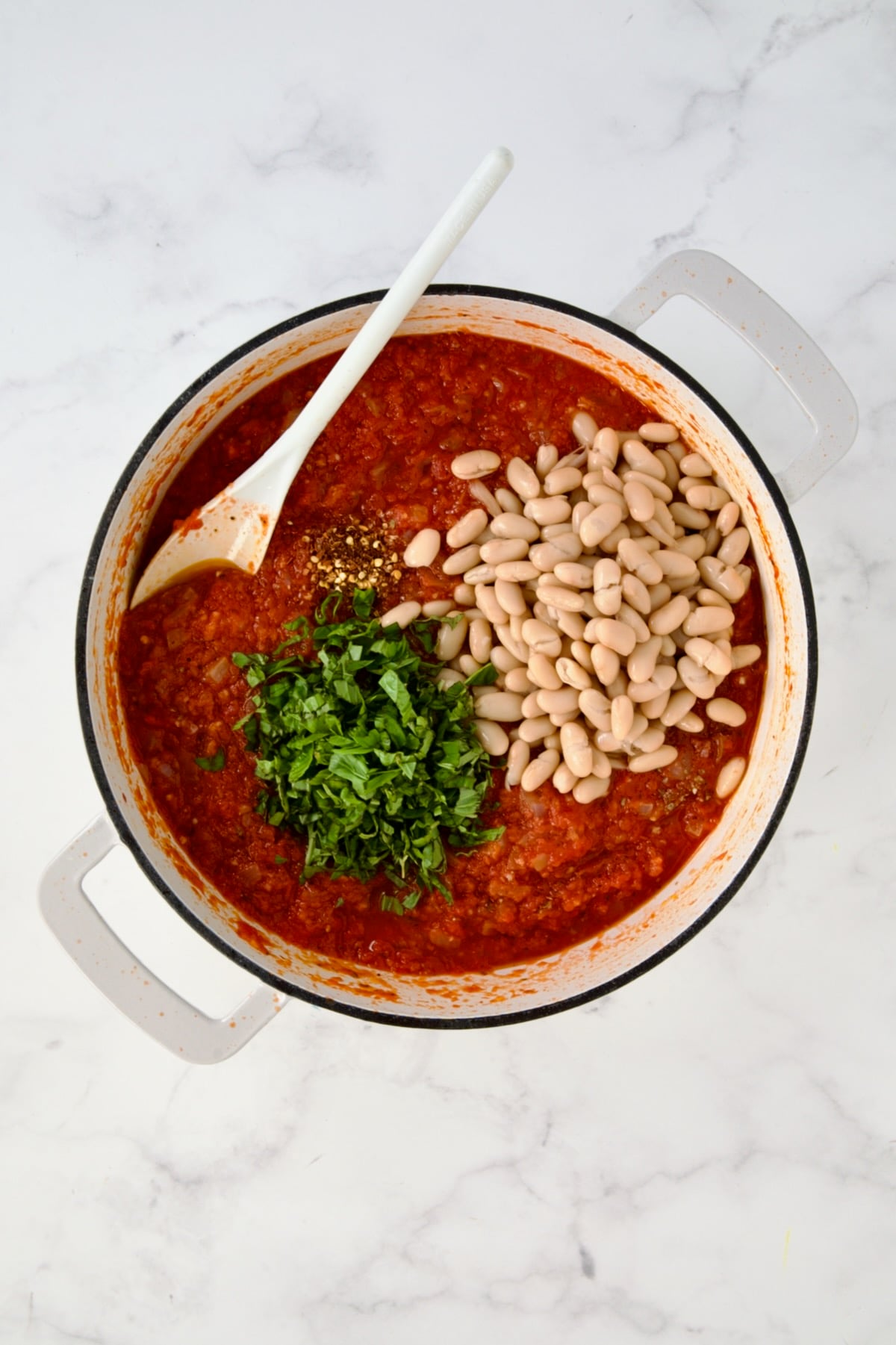 Fresh basil, beans and red pepper flakes added to tomato sauce in white enamel cast iron skillet.